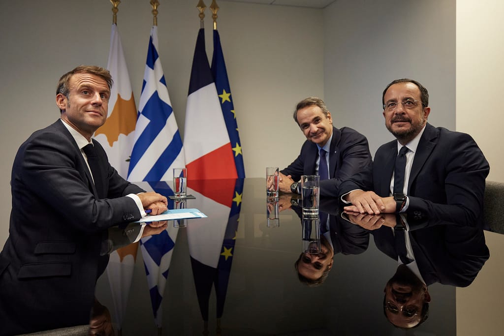 Cyprus, Greece and France officials seated at a diplomatic meeting table with national flags behind them during a trilateral discussion.