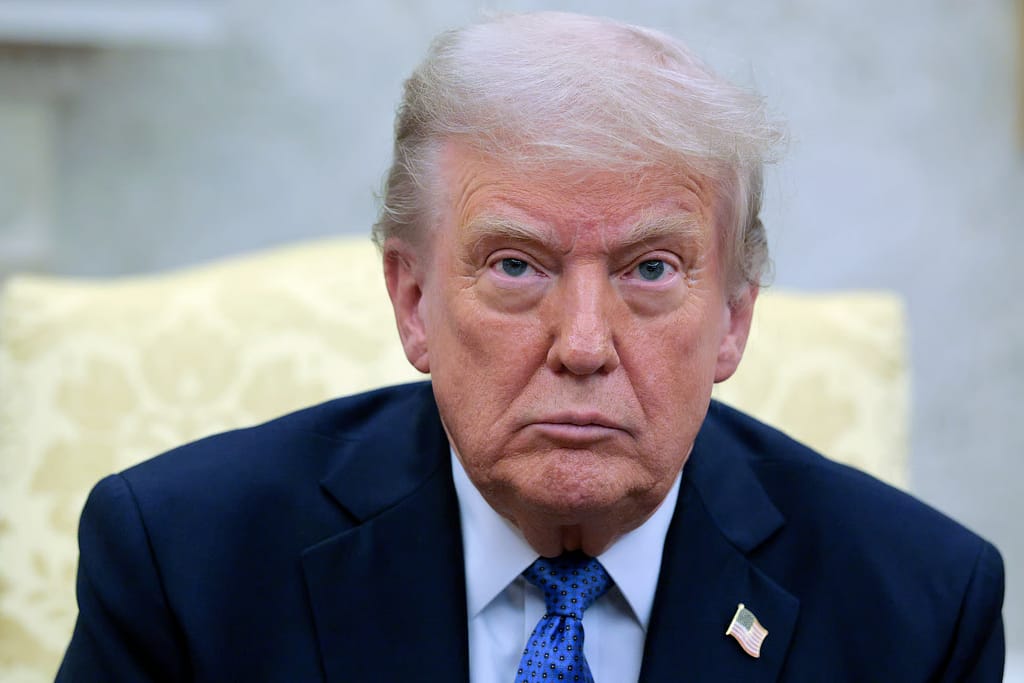 Donald Trump seated indoors during a formal interview, wearing a dark suit and blue tie, with a patterned sofa in the background