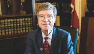 A suited official seated in an office with bookshelves and a national flag behind him during a formal interview or statement.