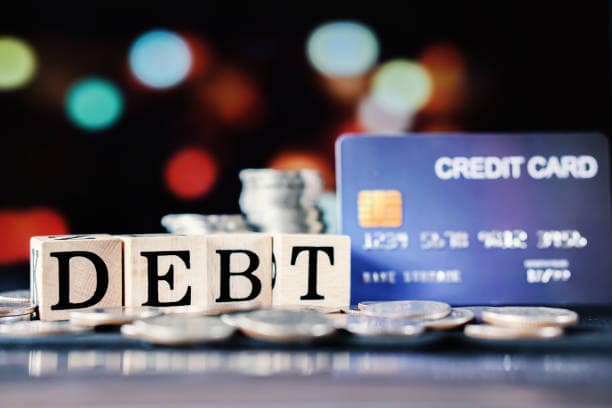 Wooden blocks spelling ‘DEBT’ surrounded by coins, with a blurred blue credit card in the background, symbolizing rising credit card debt in 2026